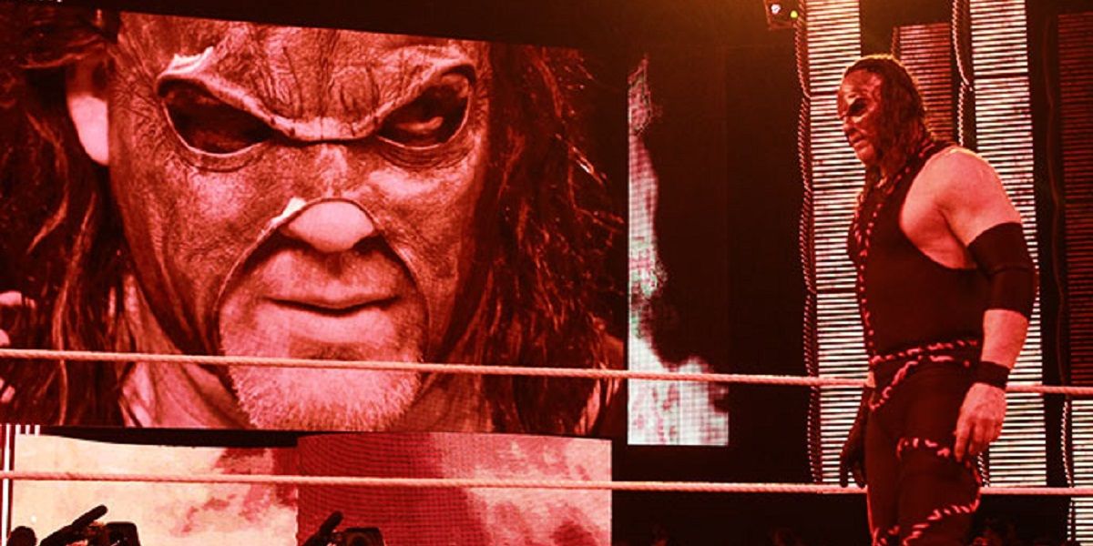 Masked Kane standing in the ring while the Titantron shows an image of him