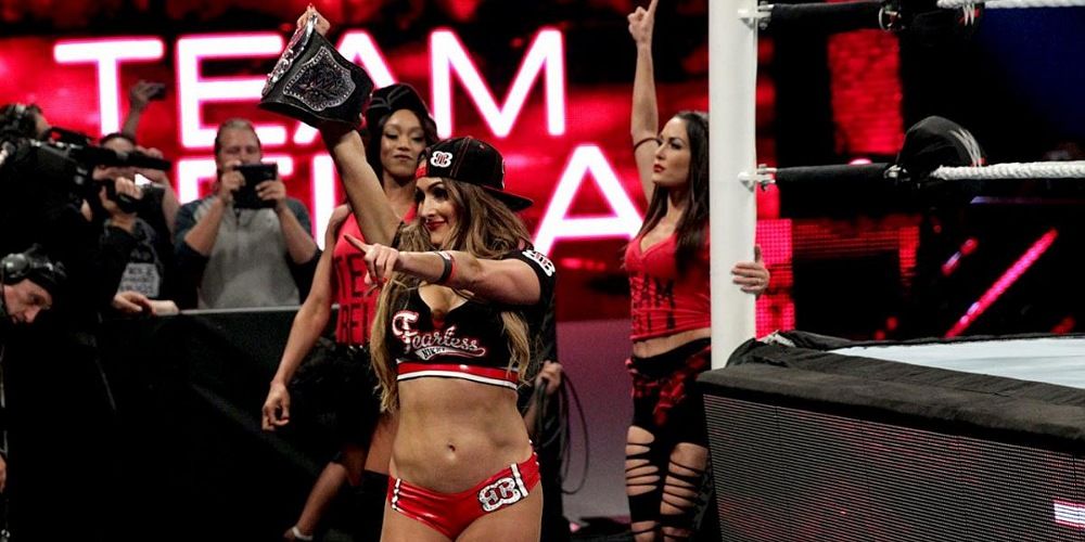 Nikki Bella holds the Divas Title and points at fans with Alicia Fox and Brie Bella behind her